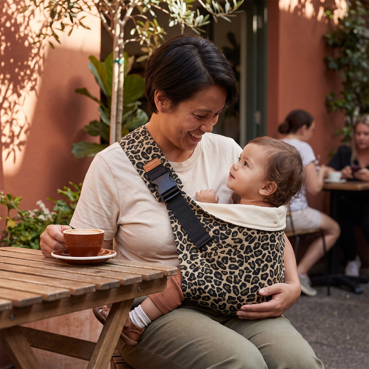 Parent carrying toddler in golden leopard baby sling at an outdoor cafe