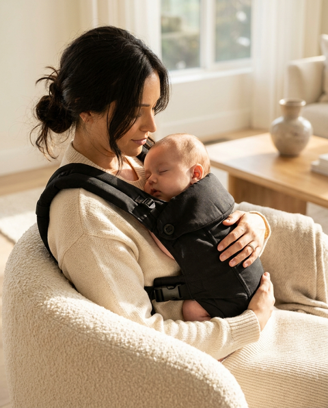 Mother wearing black ergonomic baby carrier with sleeping newborn