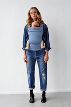Mother wearing the Née Powder Blue ergonomic baby carrier with baby in a front carry, full-length view