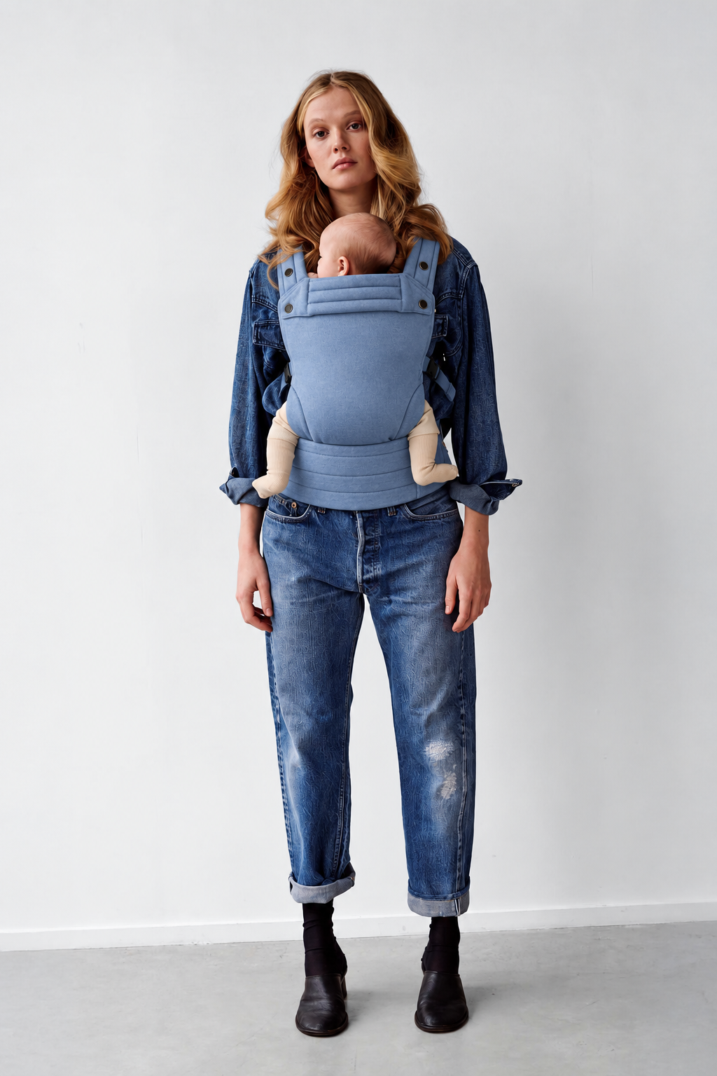 Mother wearing the Née Powder Blue ergonomic baby carrier with baby in a front carry, full-length view